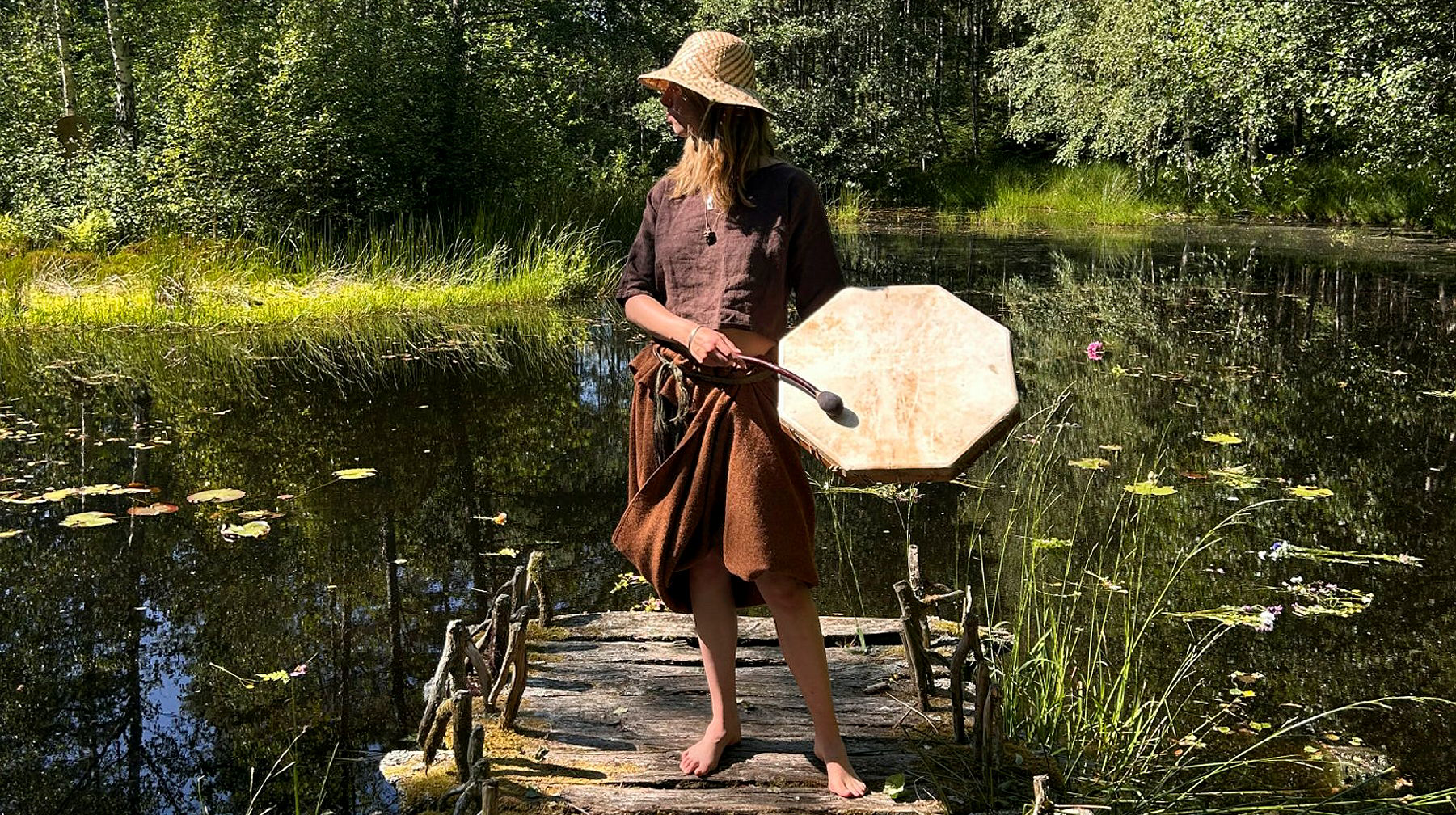 Kvinna klädd i bronsålderskläder och hatt hållandes i en trumma står på en brygga i en sjö