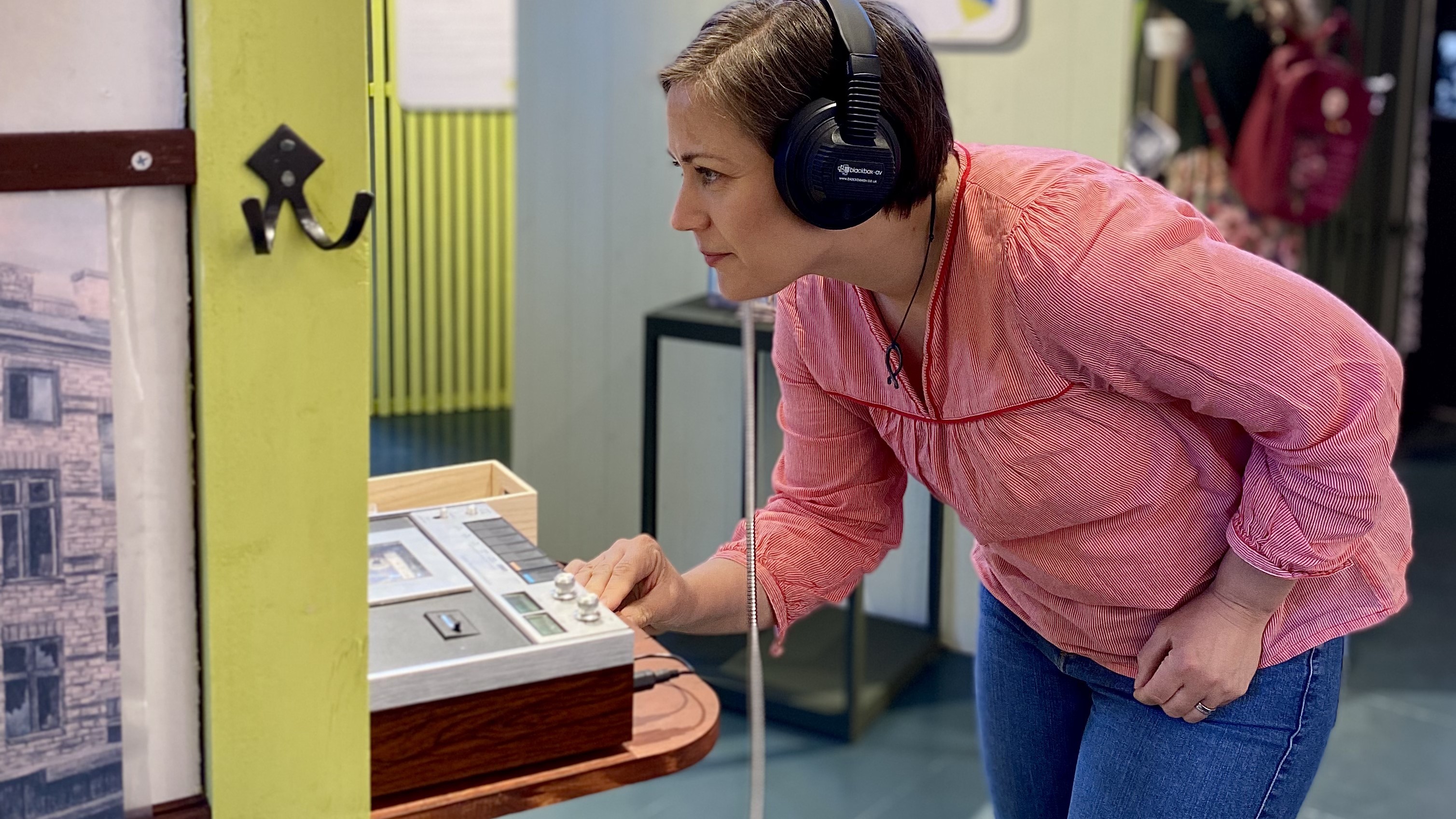 En kvinna med ett par hörlurar syns titta på något man inte ser då en vägg med en krok skymmer sikten, framför henne en bandspelare eller liknande