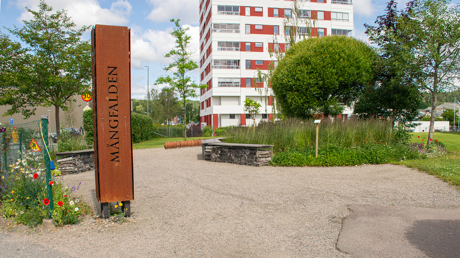 En liten grön oas har planterats i ett bostadsområde. En rund plantering med träd och perenner omges av en grusgång och bänkar. I planteringen står en liten skylt, vid ingången finns en stor metallskylt med texten "Mångfalden". 