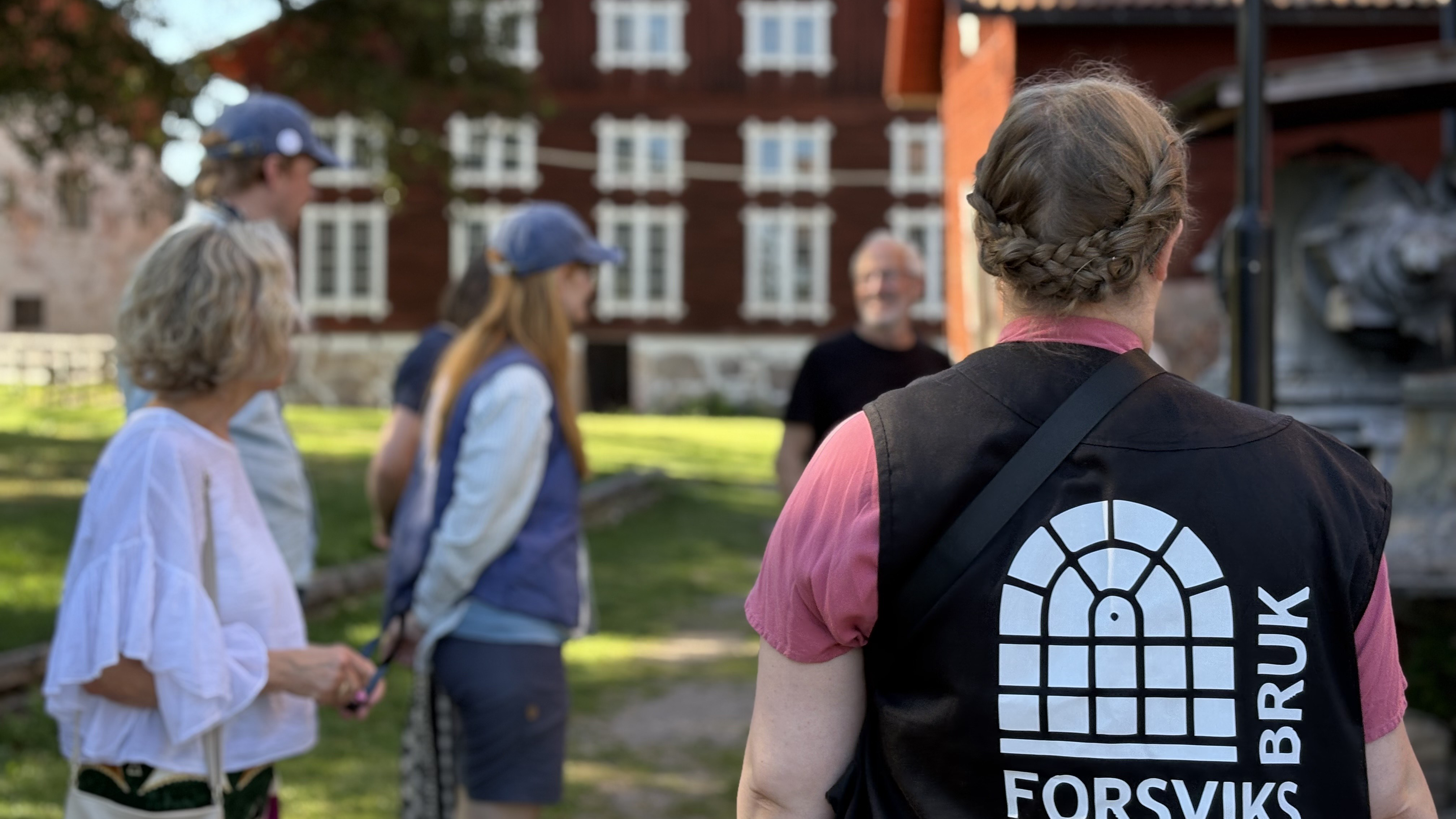 En person med guideväst med Forsviks bruks logga står vänd mot kameran, i bakgrunden en äldre stor träbyggnad samt människor