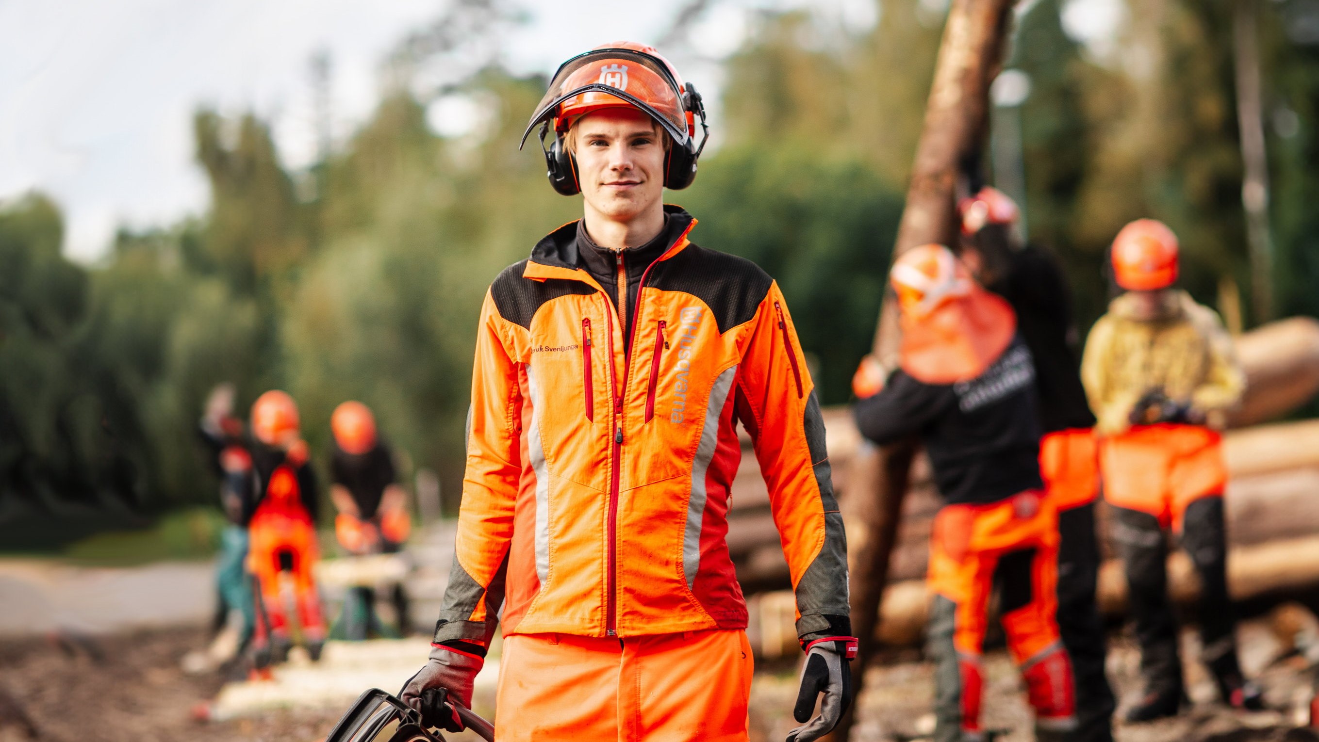 Elev med skyddskläder och hjälm håller i motorsåg med elever i bakgrunden