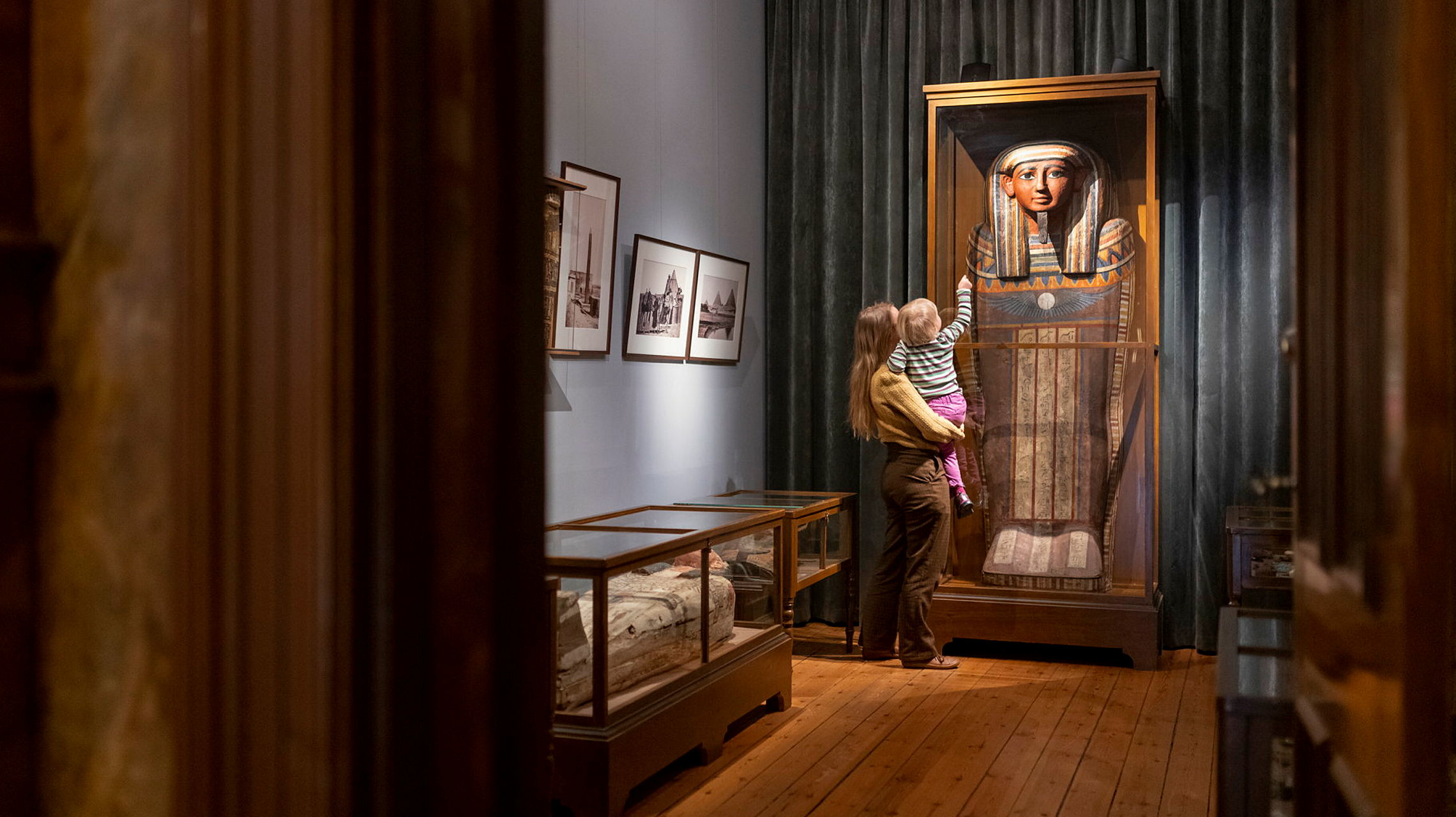 En vuxen håller ett litet barn i famnen och de tittar på en egyptisk sarkofag i en stående monter i en äldre museimiljö